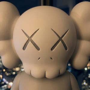 K&ouml;nnte beinhalten: Eine matte, beige Vinyl-Sammelfigur mit einem abgerundeten Kopf, ausgepr&auml;gten Ohren, X-f&ouml;rmigen Augen und einem kleinen Mund. Die Figur hat eine glatte, einfarbige Oberfl&auml;che und eine markante, stilisierte, sch&auml;delartige Gesichtsstruktur.