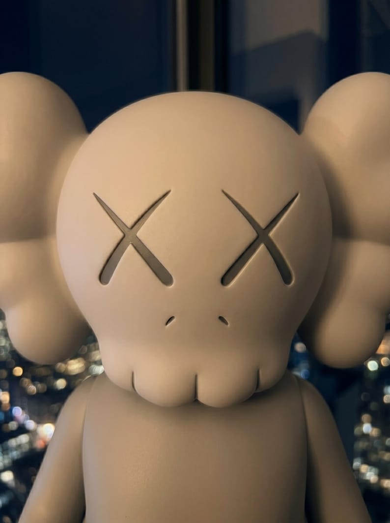 K&ouml;nnte beinhalten: Eine matte, beige Vinyl-Sammelfigur mit einem abgerundeten Kopf, ausgepr&auml;gten Ohren, X-f&ouml;rmigen Augen und einem kleinen Mund. Die Figur hat eine glatte, einfarbige Oberfl&auml;che und eine markante, stilisierte, sch&auml;delartige Gesichtsstruktur.
