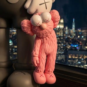 K&ouml;nnte beinhalten: Eine rosafarbene, flauschige, anthropomorphe Figur mit gro&szlig;en, runden Augen und X-f&ouml;rmigen Pupillen, die an der Hand einer gr&ouml;&szlig;eren, grauen Figur mit glatter Oberfl&auml;che h&auml;ngt. Beide Figuren haben X-f&ouml;rmige Markierungen an H&auml;nden und F&uuml;&szlig;en.