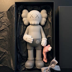 K&ouml;nnte beinhalten: Eine graue KAWS Companion-Figur mit wei&szlig;en Handschuhen und totenkopfartigem Kopf, die eine kleinere rosa Figur h&auml;lt. Das Sammlerst&uuml;ck befindet sich in einer schwarzen rechteckigen Schachtel, die mit schwarzem Seidenpapier und einer grauen Schleife ausgelegt ist.
