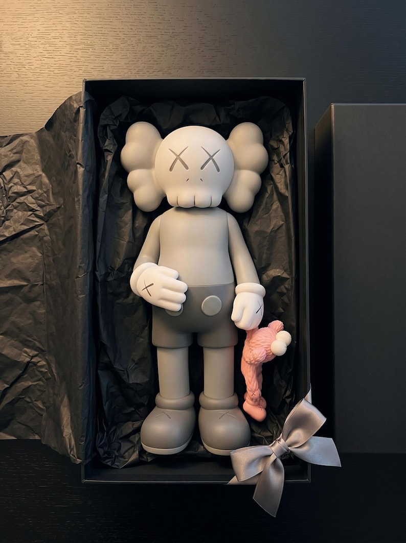 K&ouml;nnte beinhalten: Eine graue KAWS Companion-Figur mit wei&szlig;en Handschuhen und totenkopfartigem Kopf, die eine kleinere rosa Figur h&auml;lt. Das Sammlerst&uuml;ck befindet sich in einer schwarzen rechteckigen Schachtel, die mit schwarzem Seidenpapier und einer grauen Schleife ausgelegt ist.