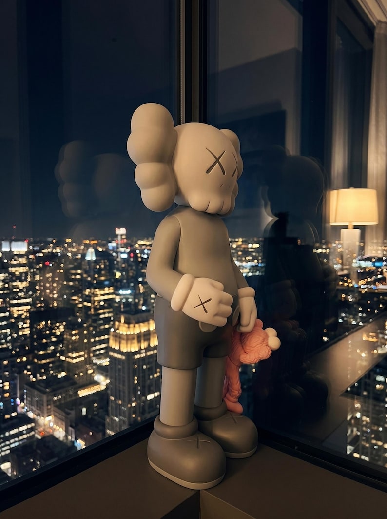 K&ouml;nnte beinhalten: Eine graue KAWS-Vinylfigur in aufrechter Position mit einem totenkopfartigen Kopf, kreuzf&ouml;rmigen Augen, abgerundeten Ohren und behandschuhten H&auml;nden mit Kreuzmarkierungen. Die Figur tr&auml;gt eine dunkelgraue Shorts und h&auml;lt eine kleine, rosafarbene Figur.