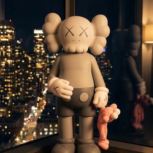 K&ouml;nnte beinhalten: Eine graue KAWS Companion Figur mit totenkopfartigem Kopf, X-f&ouml;rmigen Augen und behandschuhten H&auml;nden. Die Figur tr&auml;gt eine dunkelgraue Hose und h&auml;lt eine kleine, rosafarbene, pl&uuml;schartige Figur an der Hand mit glatten, matten Oberfl&auml;chen.