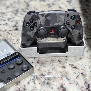 May include: A gray and black camouflage patterned video game controller with a PlayStation logo, resting in a white stand. Beside it is a silver metal tin containing a small black screwdriver and several black circular joystick thumbstick caps.