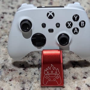 May include: A white wireless video game controller with black joysticks, a black directional pad, and black action buttons labeled A, B, X, and Y, resting on a red metal stand featuring an engraved graphic of a controller with a flame crown.