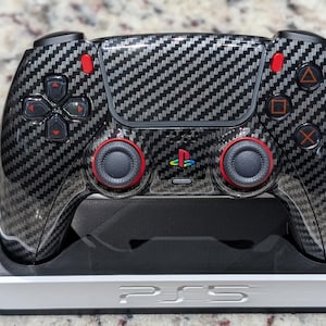Puede incluir: Un mando de videojuegos con patrón de fibra de carbono negro, detalles en rojo y un logotipo de PlayStation multicolor, apoyado sobre una base de carga blanca y negra con el texto PS5 en relieve en la parte frontal del dispositivo.