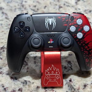 May include: Black and red PlayStation 5 controller with a Spider-Man theme, featuring a white spider logo and red accents. The controller rests on a red stand with a flame and controller graphic.