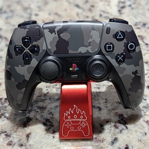 May include: A camouflage-patterned video game controller with black buttons and a red stand. The controller has a gray, black, and white camouflage design. The red stand features a flame and controller graphic.