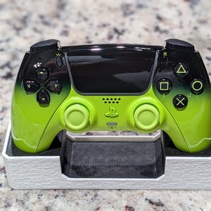 May include: A wireless video game controller featuring a lime green and black gradient finish, resting in a white textured display stand. The controller includes a central touchpad, dual analog sticks, a directional pad, and four action buttons.