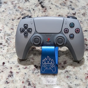May include: A gray PlayStation 5 controller resting on a blue stand. The controller has black buttons and a PlayStation logo. The stand has a graphic of a controller with flames.
