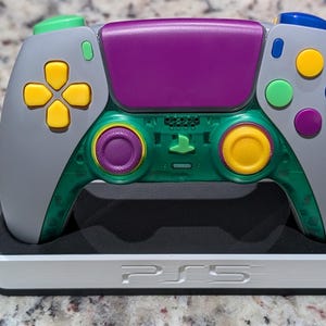 Op de afbeelding: Een aangepaste gamecontroller met een grijze behuizing, een doorschijnend groen onderste gedeelte en een paars touchpad. Het heeft gele richtingstoetsen, een groene schouderknop en blauwe, groene en paarse actieknoppen op een witte PS5-standaard.
