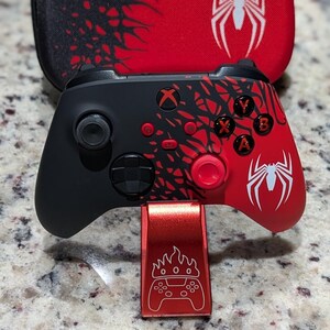 Op de afbeelding: Een zwart-rode draadloze gamecontroller met een spinnenembleem en webachtige patronen, rustend op een metallic rode standaard met een gegraveerd controller-icoon. Daarachter staat een bijpassende rood-zwarte draagtas met een wit spinnenlogo.