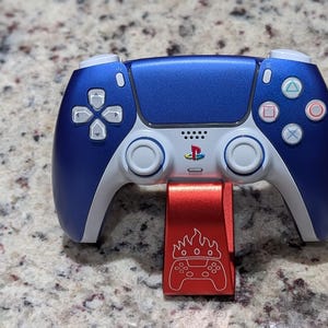 May include: A blue and white PlayStation 5 controller with a red stand. The controller has the PlayStation logo and action buttons. The stand has a flame design and a controller graphic.