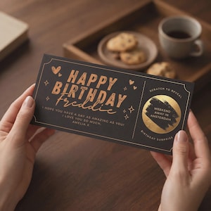 Puede incluir: Una tarjeta de cumpleaños rectangular negra con texto de color cobre que dice "HAPPY BIRTHDAY Freddie" y "I HOPE YOU HAVE A DAY AS AMAZING AS YOU! I LOVE YOU SO MUCH, AMELIA X". Incluye un área circular dorada para rascar con "SCRATCH TO REVEAL".