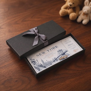 Könnte beinhalten: Eine rechteckige schwarze Geschenkbox mit einer grauen Satinschleife, die eine gedruckte Bordkarte für New York enthält. Das Ticket zeigt Illustrationen der Brooklyn Bridge, eines gelben Taxis und der Skyline der Stadt mit dem Text New York.