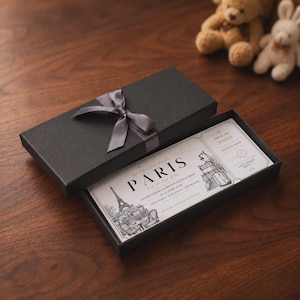 May include: A black rectangular gift box with a grey satin ribbon bow containing a printed boarding pass for Paris. The pass features an illustration of the Eiffel Tower, text reading "BOARDING PASS PARIS", "AN ANNIVERSARY ESCAPE FOR OLIVER & GRACE", and "12-15 JUNE 2028".