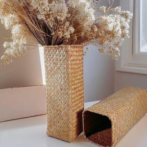 Può includere: Un vaso alto e rettangolare in vimini intrecciato contiene fiori bianchi secchi. Un secondo cesto simile è sdraiato su un lato. I cesti sono realizzati in materiale tessuto di colore naturale e poggiano su una superficie bianca.