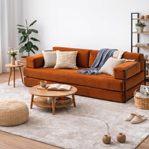 May include: A modern living room setup featuring an orange corduroy sofa with decorative pillows and a blue throw blanket. A round wooden coffee table, a woven ottoman, and a bookshelf complete the cozy space. A light gray rug covers the floor.
