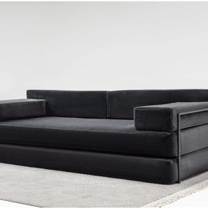 May include: A modern black corduroy sofa with a minimalist design. The sofa features a low profile, rectangular armrests, and a metal frame. It is placed on a light gray rug, creating a contrast with the dark upholstery.