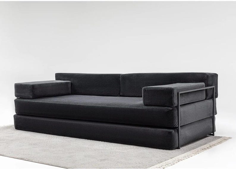 May include: A modern black corduroy sofa with a minimalist design. The sofa features a low profile, rectangular armrests, and a metal frame. It is placed on a light gray rug, creating a contrast with the dark upholstery.