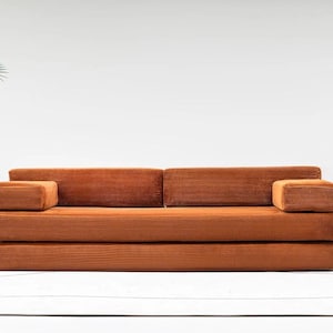 May include: A modern, corduroy-textured sofa in a warm orange hue. The sofa features a low profile with a rectangular shape, including two armrests. The design is minimalist, with a focus on clean lines and a comfortable aesthetic.