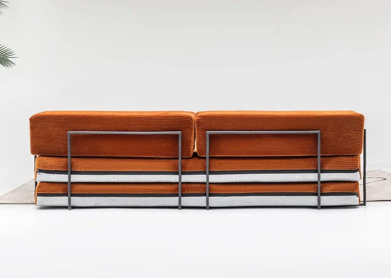 May include: An orange corduroy sofa with a metal frame. The sofa has a stacked design with layers of orange, white, and black fabric. The metal frame is dark gray.