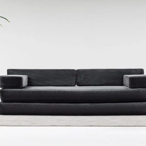 May include: A modern, dark gray sofa with a minimalist design. The sofa features a corduroy-like texture and two armrests. The sofa sits on a light gray rug. The background is a plain white wall.