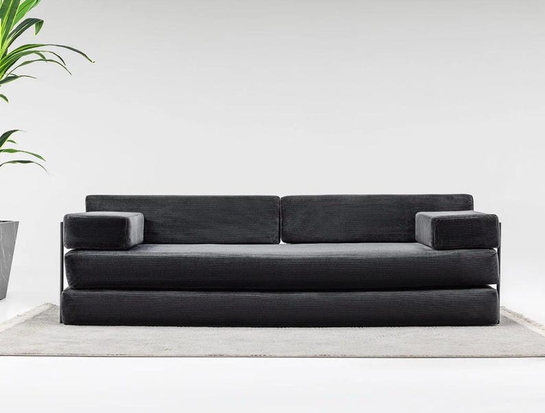 May include: A modern, dark gray sofa with a minimalist design. The sofa features a corduroy-like texture and two armrests. The sofa sits on a light gray rug. The background is a plain white wall.