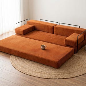 May include: A modular, rust-colored corduroy sofa with black metal frame accents. The sofa includes a matching ottoman and is placed on a round, woven jute rug. A small mug sits on the ottoman.