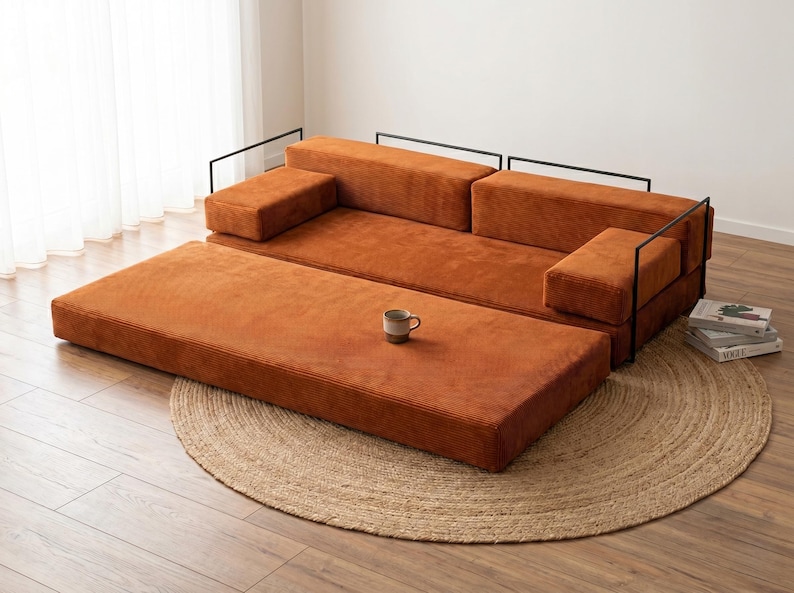 May include: A modular, rust-colored corduroy sofa with black metal frame accents. The sofa includes a matching ottoman and is placed on a round, woven jute rug. A small mug sits on the ottoman.