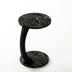Puede incluir: Una mesa auxiliar de mármol negro con tapa y base redondas, conectadas por un soporte curvo. La mesa tiene vetas blancas en todo el mármol negro. La mesa mide aproximadamente 51 cm de alto y 41 cm de diámetro.