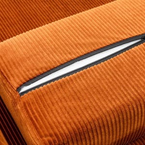 May include: Close-up of a section of orange corduroy fabric. The fabric has a ribbed texture and a black zipper is partially open, revealing a white interior. The image highlights the texture and color of the material.