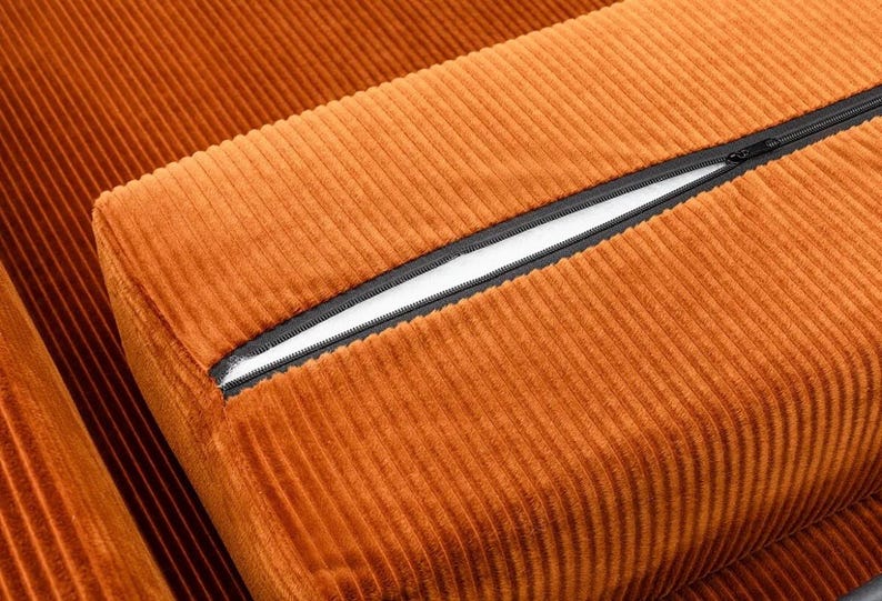 May include: Close-up of a section of orange corduroy fabric. The fabric has a ribbed texture and a black zipper is partially open, revealing a white interior. The image highlights the texture and color of the material.