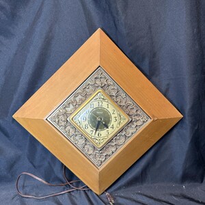 May include: A diamond-shaped wall clock with a light brown wooden frame and a central square face featuring a dark green dial, gold-tone numerals, and ornate metallic scrollwork. A black power cord hangs below, and a ruler shows 26 inches.