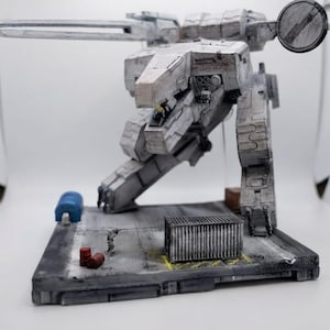 May include: A detailed gray and white mechanical robot model standing on a rectangular base. The robot features a large cannon, a circular radar dish, and articulated legs. The base includes a small shipping container, two red barrels, and a blue tank.