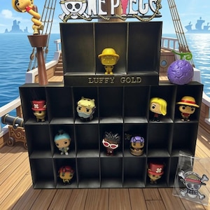 May include: A black display shelf featuring the text ONE PIECE and LUFFY GOLD, containing various small character figurines. A gold Luffy figure sits on the top shelf, while other figures occupy the lower compartments. A purple fruit and a logo patch are included.