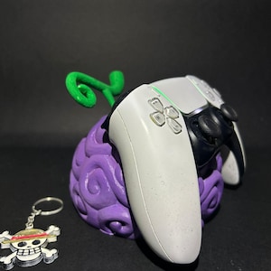 May include: A purple, textured gaming controller stand shaped like a fruit, with a green stem. A white and black gaming controller rests in the stand. A keychain with a skull and crossbones design is also present.