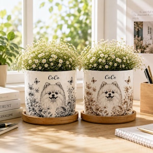 May include: Two white ceramic cylindrical planters, each featuring a black and white illustration of a Pomeranian dog, the name CoCo, and floral motifs with butterflies. Each planter sits on a circular wooden base and contains a small white flower plant.