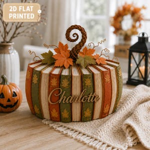 May include: A decorative pumpkin-shaped object constructed from book spines in shades of orange and green, featuring the name Charlotte in script lettering, topped with a braided stem and fabric autumn leaves, with a 2D flat printed label.