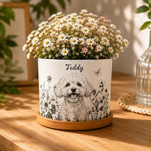 May include: A white ceramic cylindrical planter featuring a black and white sketch of a dog and the name Teddy printed above it. The planter is filled with white daisies and sits on a circular wooden saucer, showcasing floral illustrations.