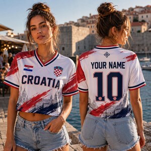 May include: A white V-neck jersey featuring red and blue brushstroke patterns, the text CROATIA, a Croatian flag, and a crest on the front. The back displays the text YOUR NAME, the number 10, and a small crest, paired with light blue denim shorts.