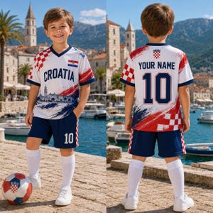Puede incluir: Un conjunto de f&uacute;tbol blanco y azul marino que incluye camiseta y pantal&oacute;n corto, con el texto CROATIA, un patr&oacute;n de cuadros rojos y blancos y un gr&aacute;fico de una ciudad costera. La espalda de la camiseta muestra YOUR NAME y el n&uacute;mero 10 en azul.