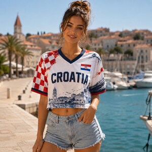 May include: A white V-neck crop top featuring a red and white checkered pattern on the right sleeve, the word CROATIA in bold blue letters, a small Croatian flag, and a blue graphic illustration of a coastal city skyline at the bottom hem.