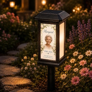 May include: A black solar-powered memorial garden light featuring a rectangular frame with a portrait of a woman named Margaret, the dates 1948-2024, and the text Your light lives on in all who knew you, surrounded by a floral border design.