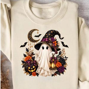 May include: A cream-coloured sweatshirt featuring a graphic of a white ghost wearing a black witch hat adorned with flowers. The ghost holds a glowing lantern next to a jack-o'-lantern, surrounded by orange and purple roses, bats, and a crescent moon.