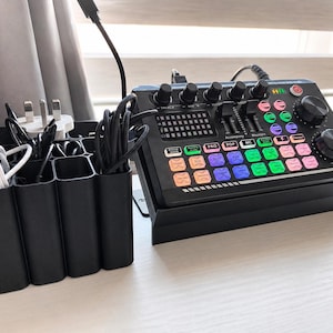 May include: Black audio mixer with multiple knobs, buttons, and a display, next to a black organizer holding various cables and plugs. The mixer has colorful illuminated buttons and a sleek design, suitable for audio recording or live streaming.
