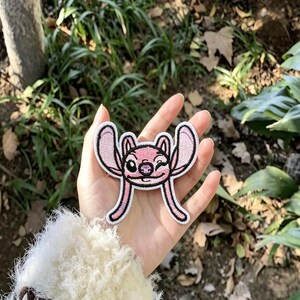 以下が含まれることがあります： 大きな耳、ウィンクした目、小さな鼻を持つピンク色のキャラクターが刺繍されたワッペン。黒い糸で縁取られ、耳と顔の内側は淡いピンク色で、下向きに伸びる2本の長く細いピンク色の突起が特徴的なデザインです。
