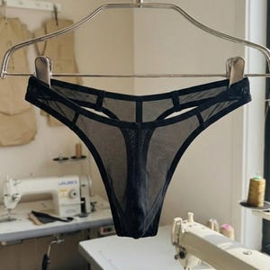 Könnte beinhalten: Ein schwarzer Tanga aus transparentem Netzstoff mit einem Riemendesign und Cut-out-Details entlang des Bundes. Das Kleidungsstück hängt an einem Metallbügel mit Clips und zeigt eine minimalistische Konstruktion mit dünnem elastischem Saum.