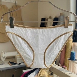 Puede incluir: Bragas blancas con ribete dorado, colgadas de una percha metálica. La ropa interior presenta un diseño delicado y texturizado. El fondo incluye equipo de costura y rollos de tela, sugiriendo una creación artesanal.