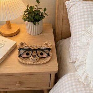 Può includere: Un portaocchiali a forma di orso all'uncinetto color beige con occhiali con montatura nera. L'orso ha guance rosa e un naso marrone. Il supporto è su un comodino di legno vicino a una lampada e una pianta in vaso.